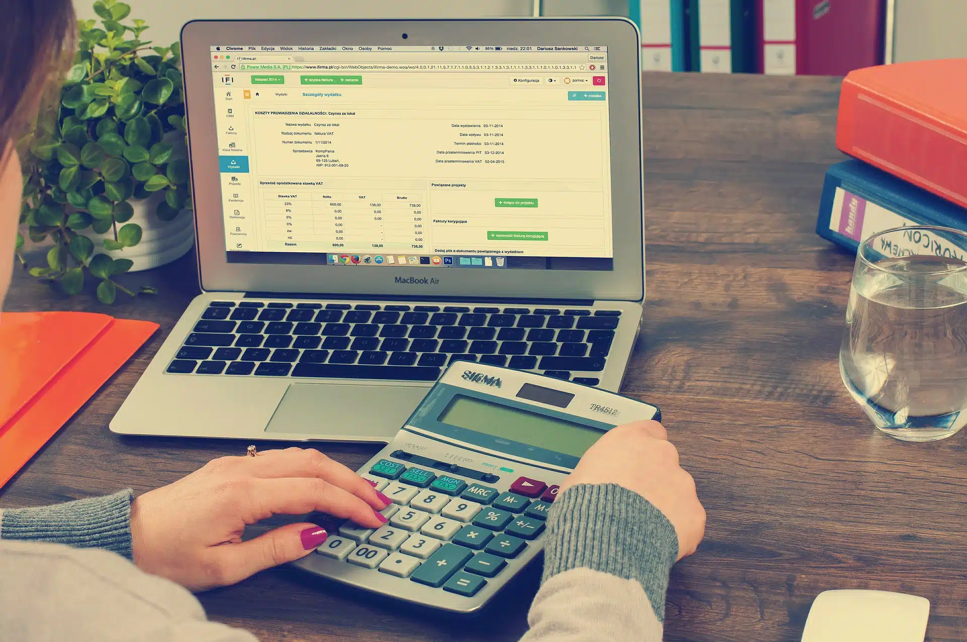 Laptop displaying financial spreadsheet, hands using calculator on wooden desk with folders, plant, glass and binders for Corporation Tax