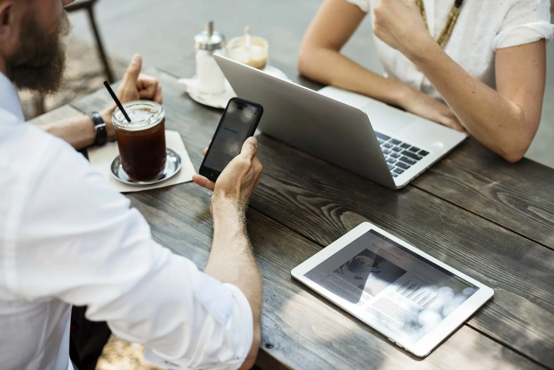 Man holding phone at table with laptop, tablet and iced drinks while talking with another person