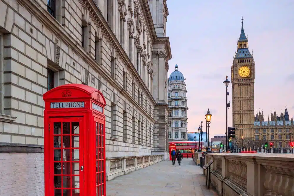 Red telephone box and red double-decker bus on a London street near Big Ben and the Houses of Parliament, representing a UK business address and director service address in England.