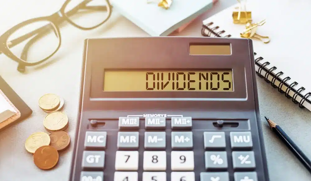 Calculator displaying the word DIVIDENDS on its screen next to coins and a notepad for calculating dividend tax