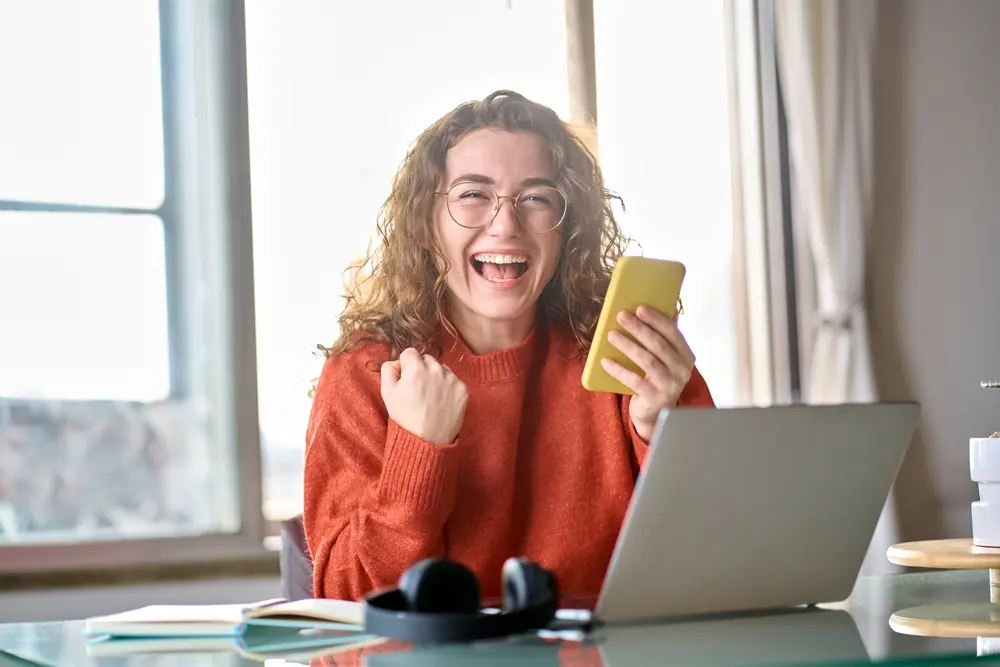 Happy woman using a phone and laptop to make money online from home.