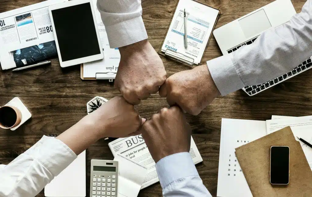 Four people bumping fists over desk with laptop, tablet, phone, calculator and documents representing business team setup