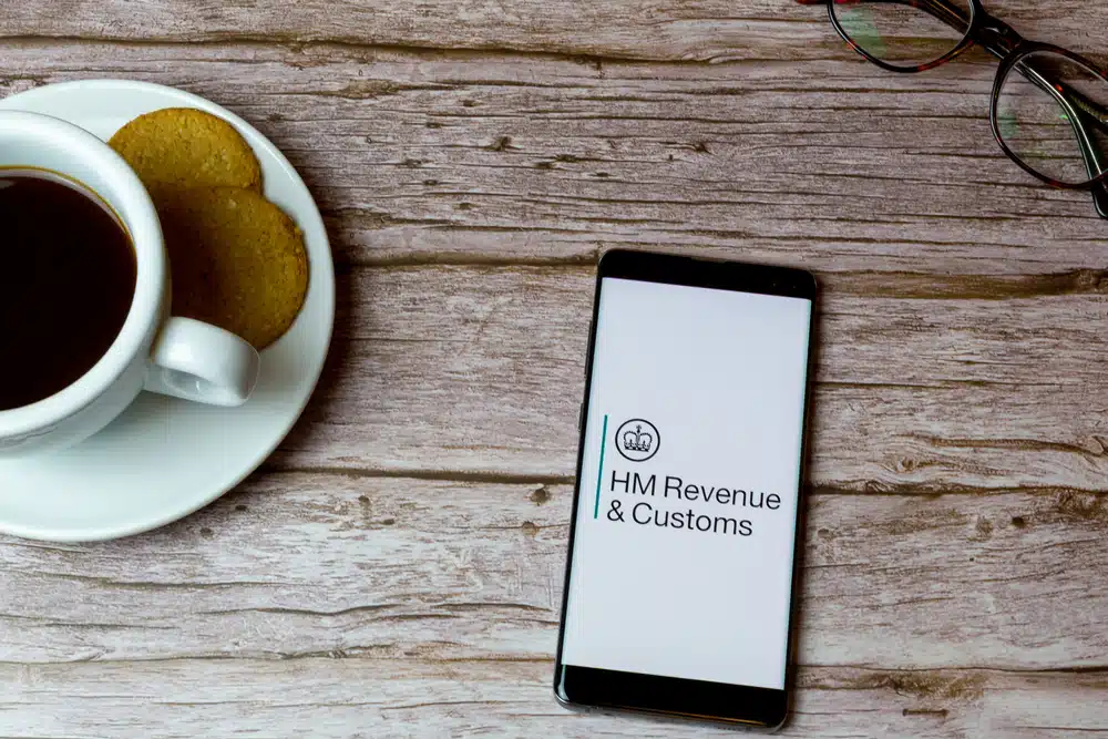 Smartphone displaying the HMRC logo on a wooden table next to a cup of tea representing UK tax brackets and HMRC tax rates