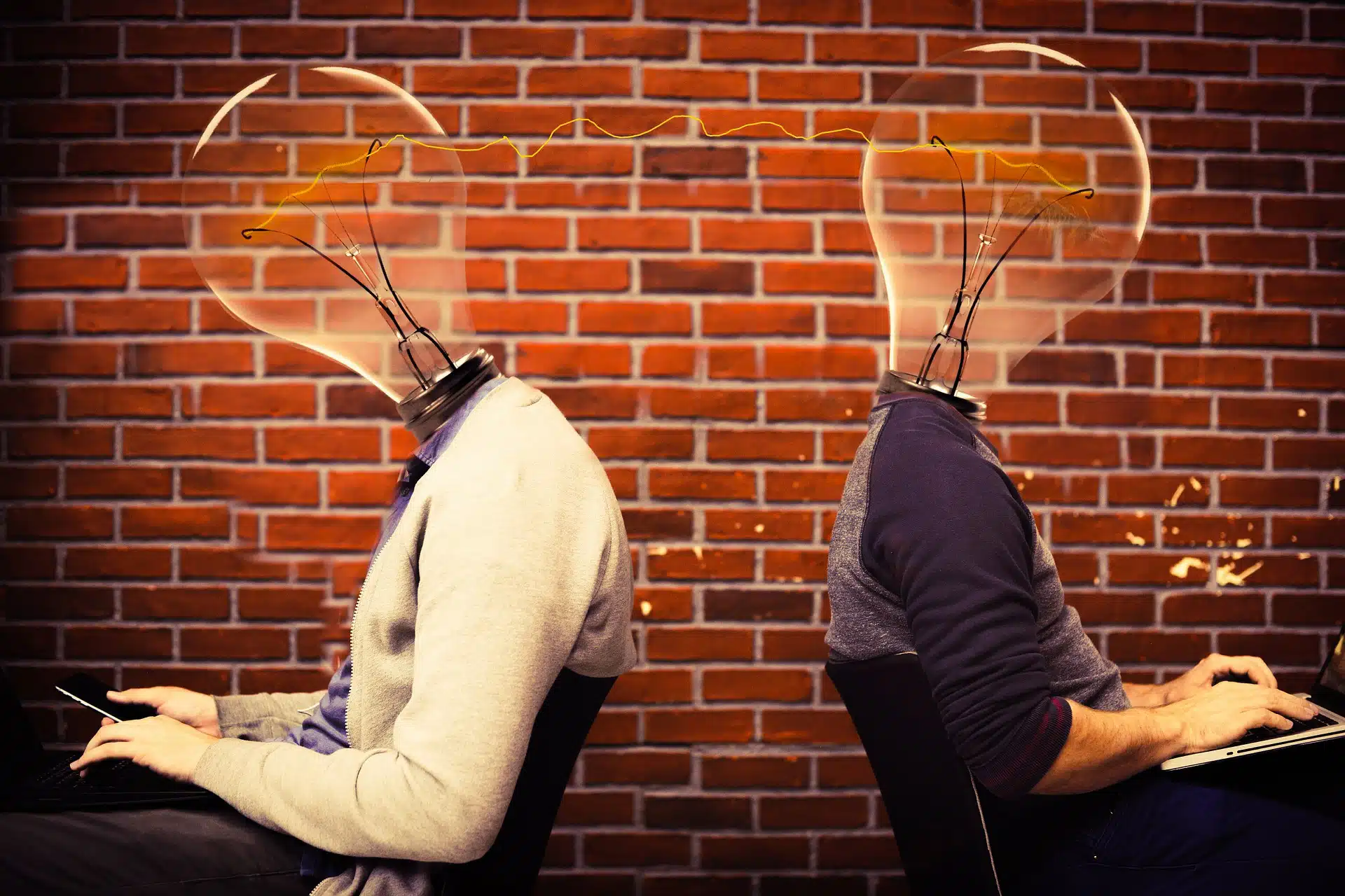 Two people with light bulb heads using laptops sitting back to back against a brick wall 