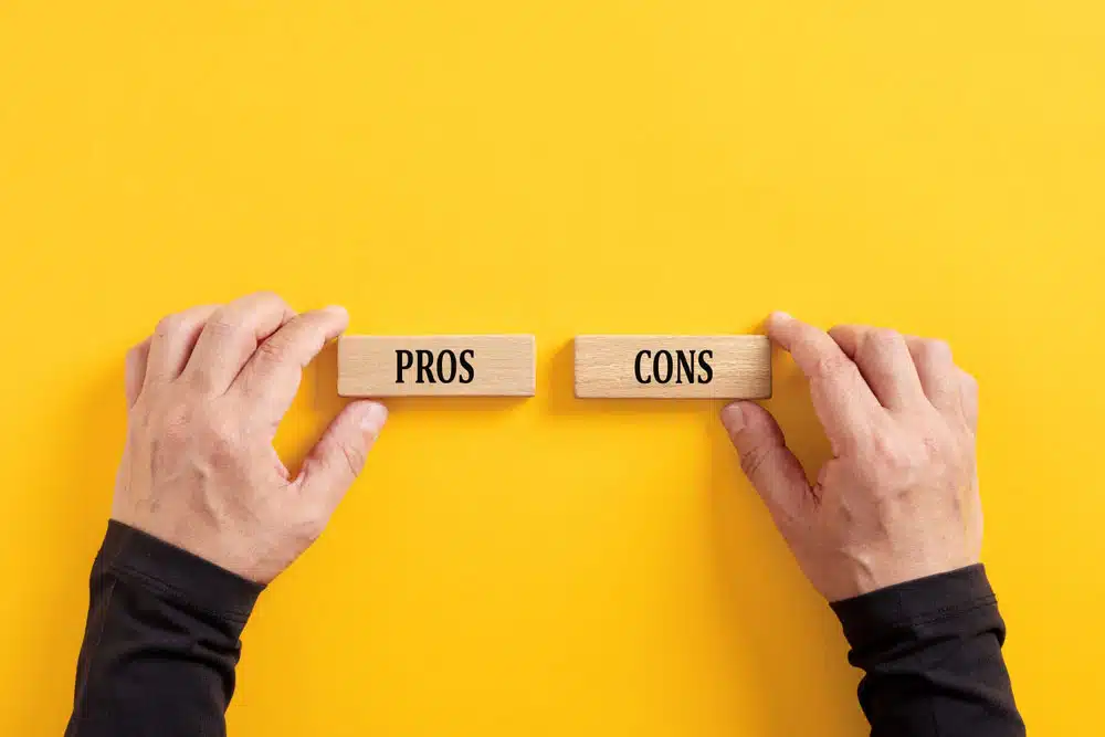 Two hands holding wooden blocks labelled Pros and Cons against a yellow background, representing the advantages and disadvantages of voluntary VAT registration for UK businesses.