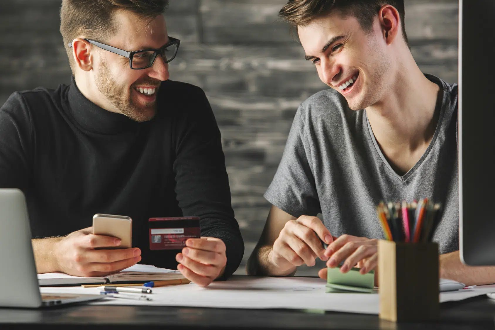 Two men smiling at desk holding bank card and phone beside laptop, papers and pens illustrating business credit score improvement